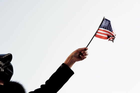 A-woman-participating-in-a-protest-in-Washington-DC-holding-a-burning-US-flag-image-by-Marco-Verch-via-Flickr-CC-e1618999488384.jpg