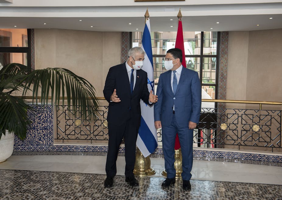 Two men wearing suits and COVID-19 masks chat with their backs to the flag of Israel on the left, and that of Morocco to the right.