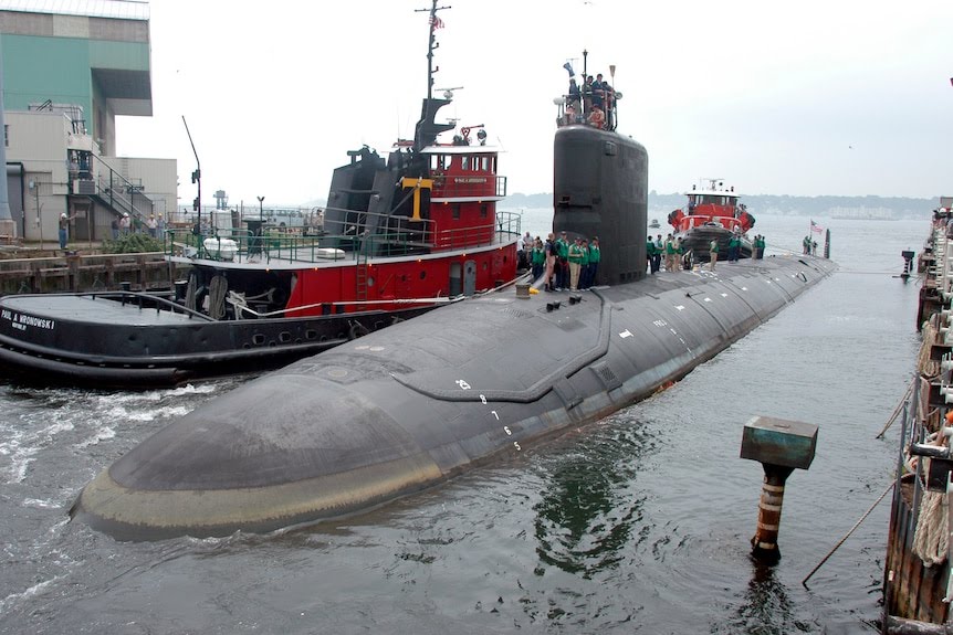 A surfaced nuclear submarine in dock next to a tug boat. 