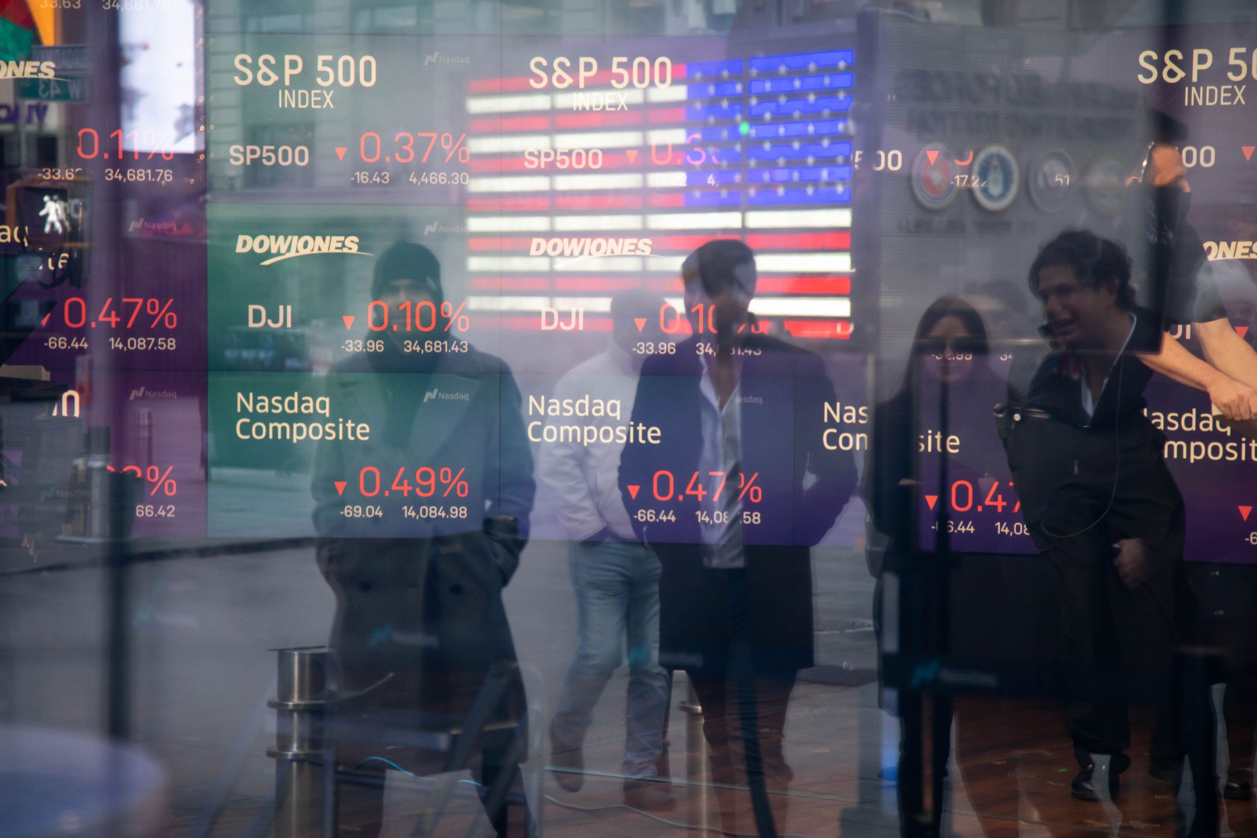 People walking by a reflection of monitors displaying stockmarket information.