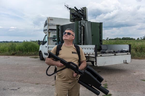 A man standing in a brown outfit, holding a large black device that looks like a plastic gun, staring up to the sky with a truck and a field behind him.