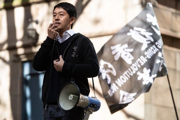 A man speaks into a mic attached to a megaphone, as a black and white flag waves behind him.
