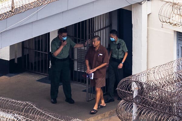A man in a brown shirt and shorts is led through a prison exit by two men in green shirts and pants.