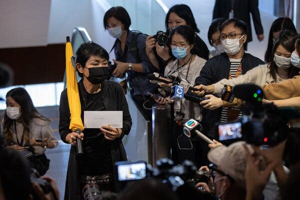Journalists point microphones at a woman in black holding a yellow umbrella.