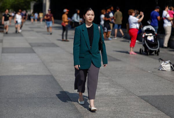 A woman in a green blazer, black blouse and gray pants strolls along a gray walkway.